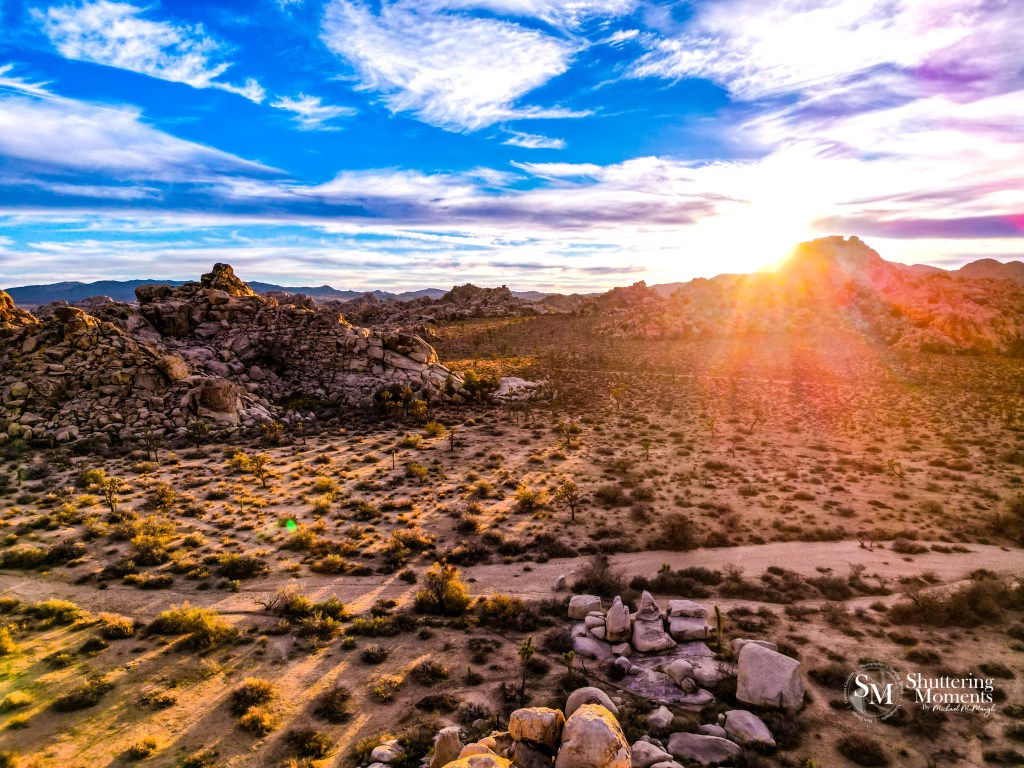 A breathtaking landscape of Joshua Tree National Park at sunset, captured by Michael McMaugh of Shuttering Moments. The image showcases rugged rock formations, iconic twisted Joshua trees, and a sky painted with warm hues of orange, pink, and purple. Professionally edited to enhance the rich textures and colors, this photograph highlights the vastness and beauty of this stunning desert location."
Let me know if you need any refinements!