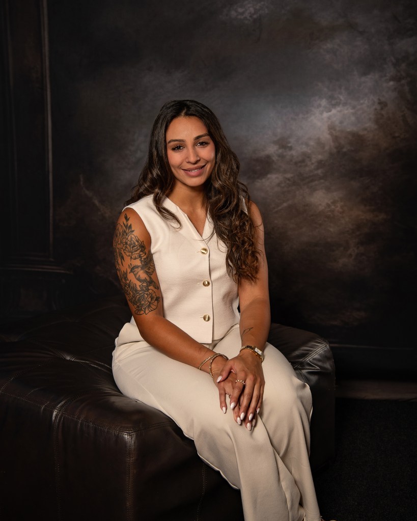 Ana Sanchez seated with her hands in her lap wearing a cream vest and matching cream pants in a professional studio portrait.