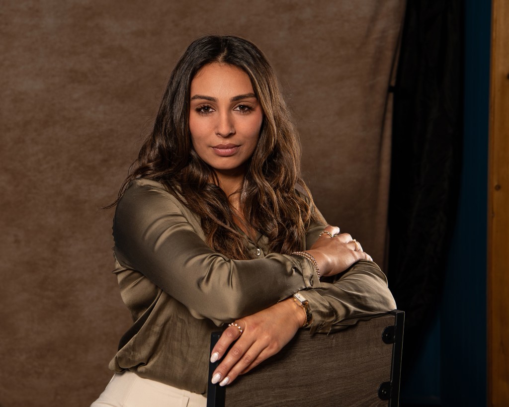 Ana Sanchez seated behind a backwards studio chair with her arms crossed, wearing an olive green silk shirt and cream pants.