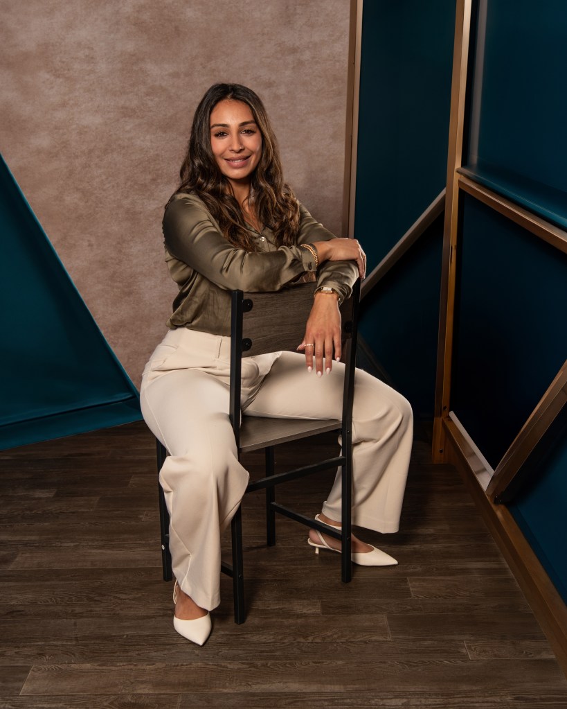 Ana Sanchez standing in an olive green silk shirt and cream pants against an abstract studio background for a full-body realtor portrait.