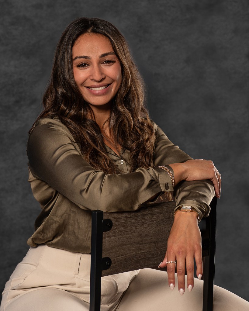 Ana Sanchez seated in an olive green silk shirt and cream pants against a blue studio backdrop during a professional branding session.