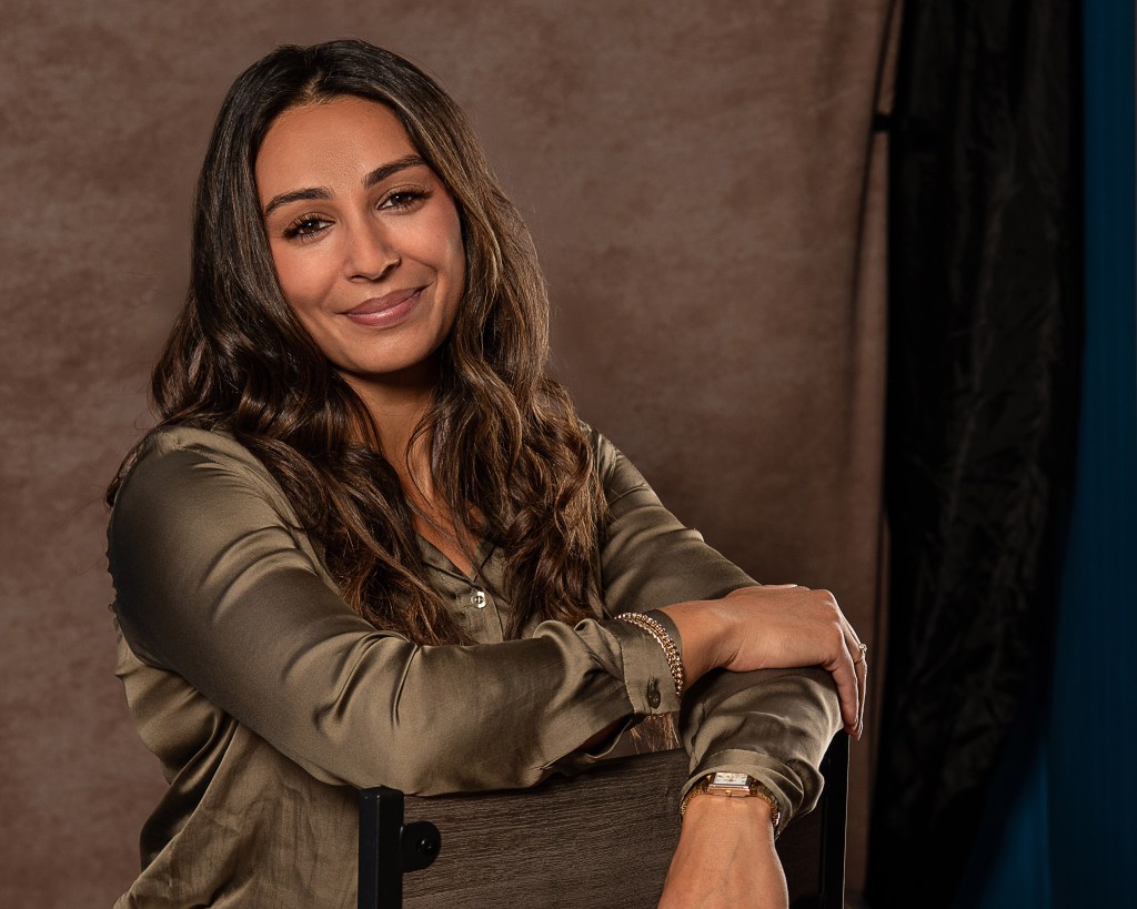 Ana Sanchez leaning casually against a backwards studio chair while wearing an olive green silk shirt and cream pants.
