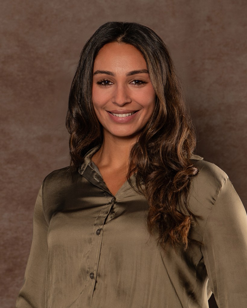 Ana Sanchez posing beside a backwards studio chair with one arm resting on the chair, wearing an olive green silk shirt and cream pants.